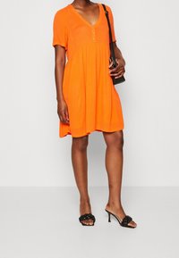 Kurzes orangefarbenes Kleid mit V-Ausschnitt, kurzen Ärmeln und Knopfdetails. Trägt man mit schwarzen geflochtenen Sandalen mit Absatz. Einfaches, fließendes Design.