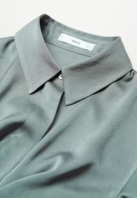 Chemise grise douce à boutons, col pointu, texture lisse et étiquette blanche visible à l’intérieur du col portant la mention MNG.
