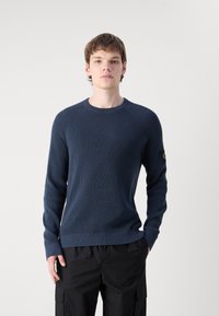 Maglione a maglia blu navy con texture a coste, scollo rotondo e maniche lunghe. Presenta un'etichetta con il logo sulla manica sinistra. Abbinato a pantaloni scuri.
