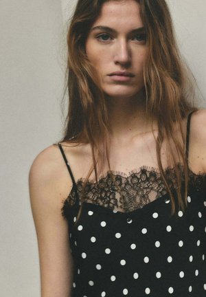 Joven mujer con cabello largo y castaño, vestida con un vestido negro de lunares con detalles de encaje, expresión neutral, sobre un fondo sencillo.