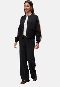 Black bomber jacket with peach-colored shoulder stripes, snap buttons, and a front pocket, paired with loose black trousers and black footwear.