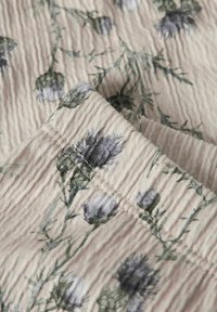 Tissu floral beige avec des ondulations texturées. Présente des fleurs vertes et bleues avec des tiges, mettant en avant un motif subtil rappelant des plis.