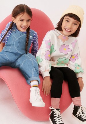 Dve nasmejani dekleti sedita na rožnati ukrivljeni stol, ena v modrih kombinezonih in črtasti majici, druga v cvetlični majici, črnih pajkicah in rumeni kapici.