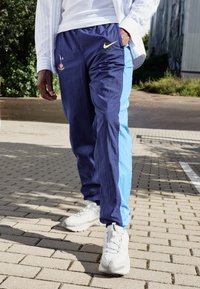 Marineblaues und hellblaues Jogginghose mit elastischem Bund, ausgestattet mit einem Nike-Logo und gestickten Akzenten, kombiniert mit weißen Sportschuhen.