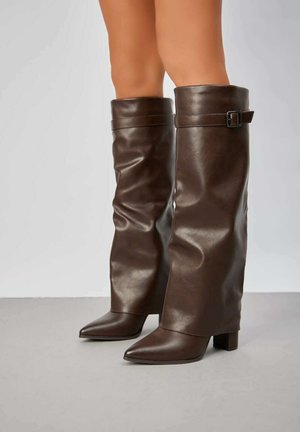 Bottes en cuir marron montantes avec un design décontracté, bout pointu et talon carré. Présente une sangle avec une boucle en détail sur le dessus.