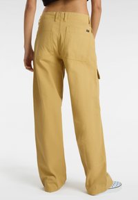 Pantaloni cargo a gamba larga in un tessuto giallo senape. Presentano due tasche posteriori e tasche cargo laterali. Tessuto liscio e materiale resistente.