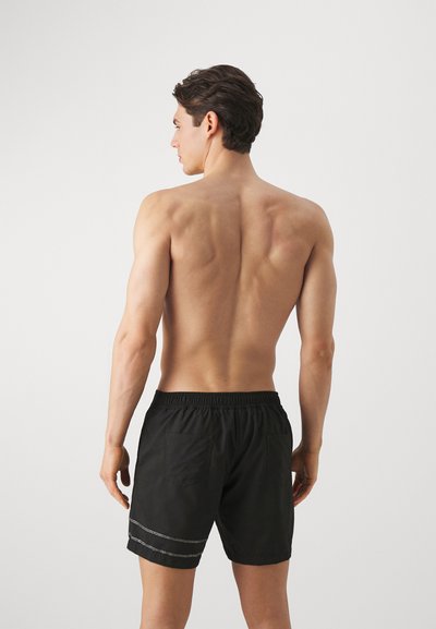 Sorte svømmeshorts med elastisk liv, med to baklommer og hvite horisontale sømmer. Utsikt bakfra.