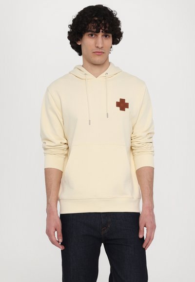 Cream hooded sweatshirt featuring a brown cross patch on the left chest, drawstrings, and a front kangaroo pocket. Cotton blend material.