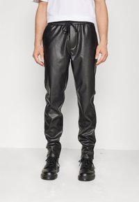 Schwarze Jogginghosen aus Kunstleder mit elastischem Bund und Kordelzug. Sie haben eine glatte Textur und schmal zulaufende Beine. Kombiniert mit schwarzen Stiefeln.