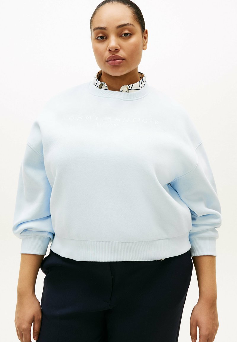 Kvinde med trukket hår, iført lyseblå Tommy Hilfiger-sweatshirt over mønstret skjorte med krave og sorte bukser, stående mod en hvid baggrund.