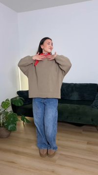 Beiger oversized Pullover mit gerippten Bündchen, kombiniert mit hellblauen Jeans und braunen Schuhen. Roter Schal betont den Halsausschnitt. Innenaufnahme.