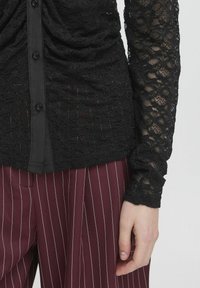 Top in pizzo nero con maniche lunghe, dotato di chiusura a bottone e motivo testurizzato. Abbinato a pantaloni a righe bordeaux.