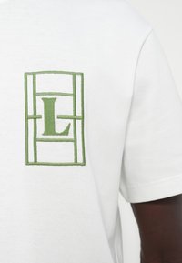 Λευκό βαμβακερό μπλουζάκι με κοντά μανίκια και στρογγυλή λαιμόκοψη, διακοσμημένο με πράσινο κεντητό τετράγωνο σχέδιο που περιλαμβάνει το γράμμα «L».