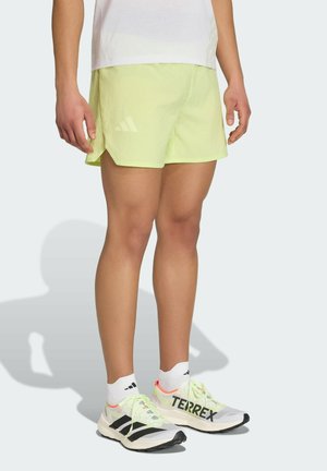 Person iført lysegule Adidas shorts, hvide sokker og hvide Adidas Terrex løbesko står på en lys baggrund.