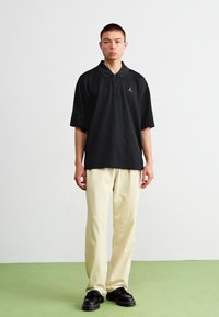 Μαύρο πόλο μπλουζάκι με κοντά μανίκια και γιακά, που διαθέτει ένα μικρό λευκό λογότυπο. Φοριέται με ανοιχτό μπεζ παντελόνι και μαύρα παπούτσια. Απλό φόντο.