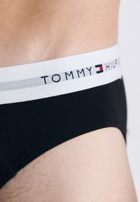 Nærbillede af talje og hofte på en person iført sorte underbukser med hvid talje, der har Tommy Hilfiger-logo og flagemblem.