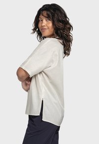 Cream short-sleeved shirt with a relaxed fit, featuring side slits and a soft texture. Worn over dark pants, shown in a side view.