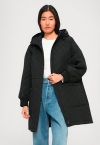 Jonge vrouw in een zwart gewatteerd hoodie-jasje, witte shirt en blauwe high-waisted jeans, staand tegen een effen lichte achtergrond.