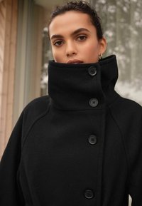 Manteau en laine noir avec un col montant, doté de grands boutons noirs et d'une surface texturée, conçu avec des manches raglan pour un ajustement décontracté.