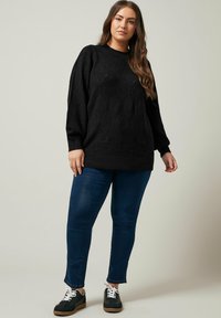 Maglione nero lavorato a maglia con un motivo a diamante strutturato, abbinato a jeans slim blu scuro e sneaker nere con lacci bianchi e suola marrone.