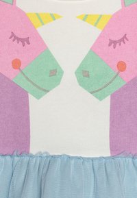 Tričko s motivem jednorožce, které má pastelové růžové a zelené grafiky na bílém pozadí; obsahuje světle modré tylové volány na spodní části.