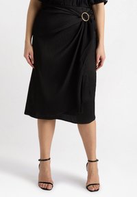 Zwarte knielange wikkelrok met geplooide textuur en een gouden ringdetail in de taille. Gedragen met zwarte sandaaltjes met banden.