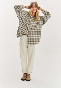 Kobieta poprawiająca brązową dzianinową czapkę, ubrana w oversize'ową koszulę w beżowo-brązowo-szara kratę, kremowe spodnie i beżowe buty trekkingowe z różowymi sznurowadłami.