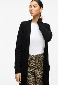 Lång svart kofta i mjukt material, öppen fram, ribbade ärmslut och två framfickor, kombinerad med en vit skjorta och leopardmönstrade byxor.