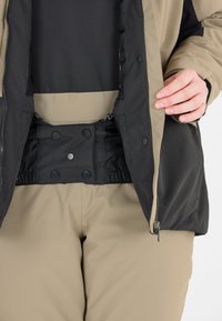 Giacca da esterno beige con accenti neri, dotata di chiusura a zip, bottoni automatici in vita e tessuto testurizzato. Abbinata a pantaloni beige.