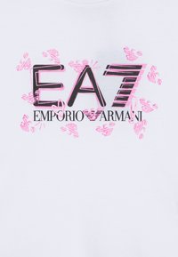 Fehér pamut póló, amelyen egy nagy rózsaszín és fekete logó grafika látható, rajta az "EA7 EMPORIO ARMANI" felirattal és játékos dizájnelemekkel körülvéve.