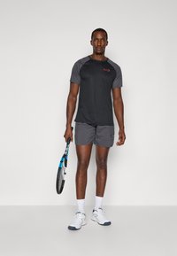 Ensemble de vêtements de sport comprenant un t-shirt noir à manches courtes avec des accents gris et un logo rouge, associé à un short gris. Tenant une raquette de tennis.