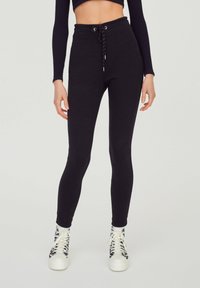 Black high-waisted leggings with a drawstring waistband and tight fit, made from a smooth, stretchy material, paired with patterned sneakers.