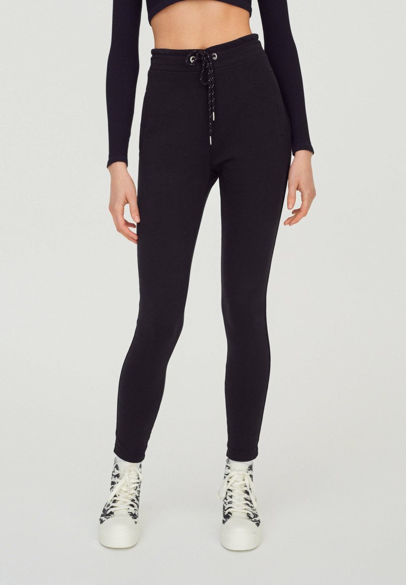 Black high-waisted leggings with a drawstring waistband and tight fit, made from a smooth, stretchy material, paired with patterned sneakers.