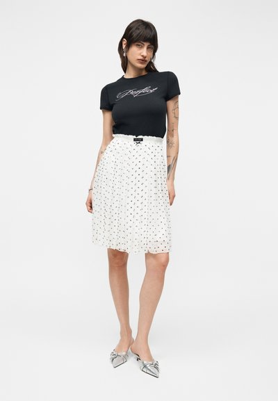 Γυναίκα με σκούρα μαλλιά που φοράει μαύρο μπλουζάκι με τη λέξη "perfect", φούστα με λευκά πουά και μαύρο φιόγκο, και ασημί μυτερά παπούτσια.