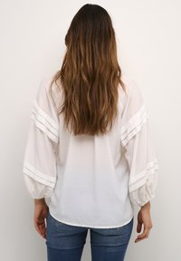 Blusa bianca con maniche voluminose e righe verticali. Tessuto leggero, chiusura a bottoni sul retro e orlo curvato. Indossata con jeans blu.
