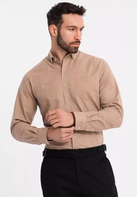 Chemise à manches longues beige en tissu lisse avec un col boutonné et deux poches poitrine, associée à un pantalon noir.