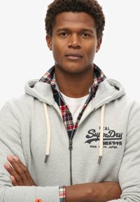 Sudadera gris con cremallera y cordones, con un logo impreso. Debajo, es visible el cuello de una camisa de cuadros en rojo y azul. Textura de tela suave.