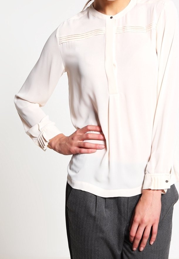 Blusa beige chiaro con scollatura rotonda, pieghe verticali sul petto, maniche con polsini e dettaglio con bottone. Tessuto morbido e liscio con una vestibilità comoda.