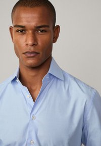 Camicia a scacchi blu chiaro con colletto button-down, caratterizzata da una sottile texture e bottoni in perla, indossata da un modello su uno sfondo neutro.