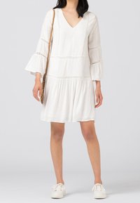 Vit klänning med utsvängda ärmar, V-ringning och utskärningar. Mjuk tyg, lager-på-lager kjol, och matchad med vita sneakers.