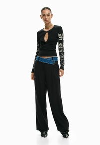 Zwart top met lange mouwen en een keyhole-uitstulping met witte tekstprint; gecombineerd met zwarte wijde broekspijpen en een blauwe spijkerbroekriem als accent.