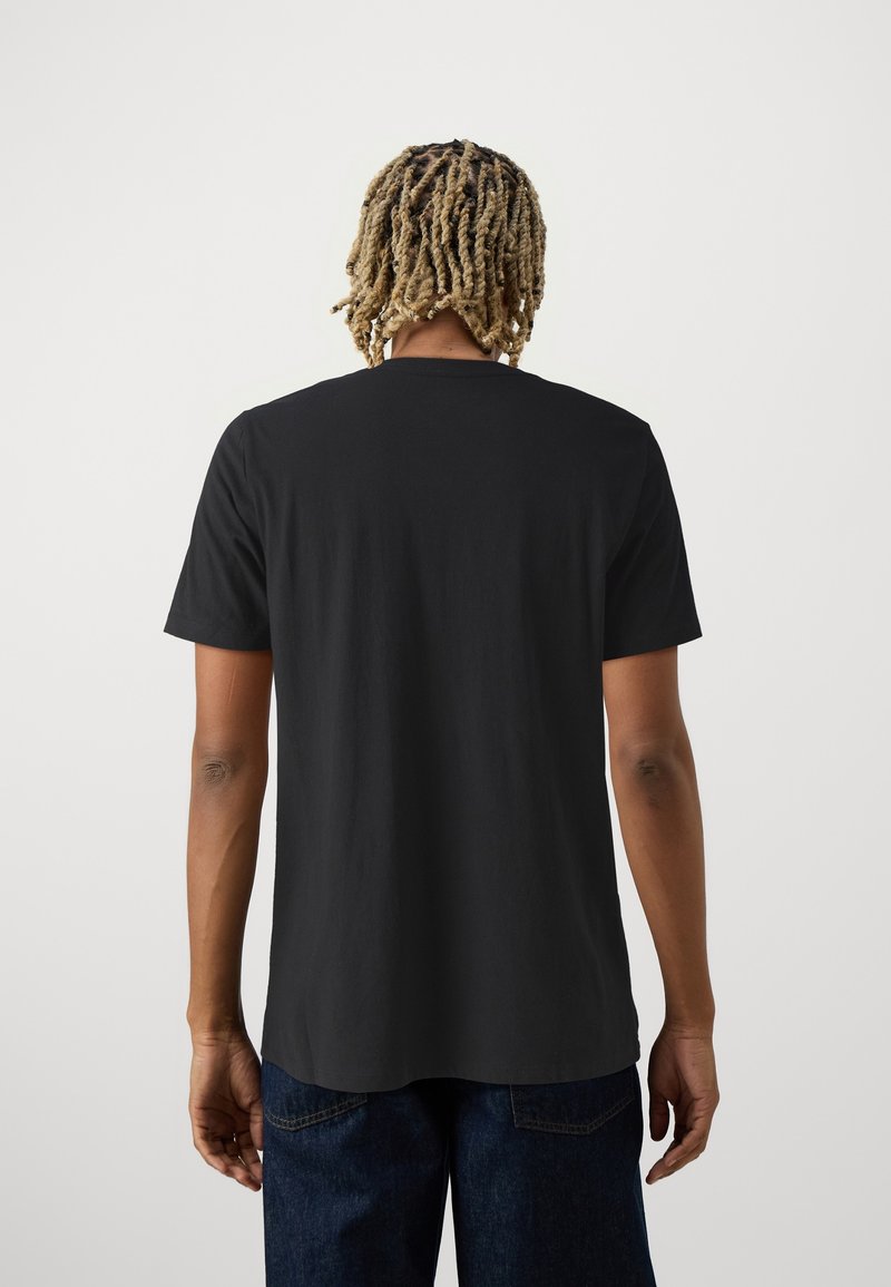 Homme aux dreadlocks blonds se tient dos à la caméra, portant un t-shirt noir uni à manches courtes et un jean bleu foncé sur fond blanc.