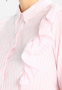 Blusa a righe rosa chiaro con bordo arricciato lungo la spalla, chiusura a bottoni e design colletto classico. Materiale in cotone morbido.