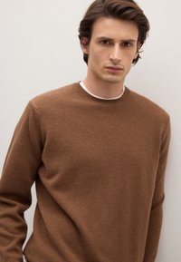 Pull marron à col rond en matière douce et texturée avec col côtelé, coupe ajustée et manches longues. Porté sur une chemise blanche.