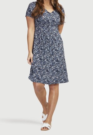 Kvinde iført en knælængde marineblå kjole med hvidt og lyseblåt blomsterprint og hvide sandaletter med åbne tæer, stående mod en lys baggrund.