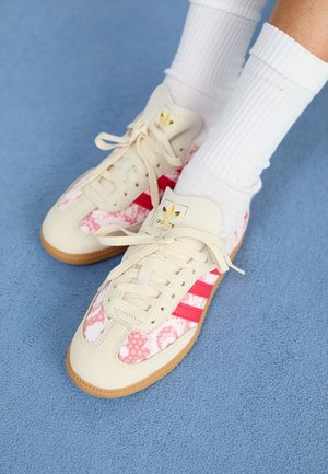 Crème-kleurige sneaker met roze bloemenaccenten, rode strepen en beige veters. Beschikt over een gestructureerde gumrubberen zool en een gouden logo op de tong.