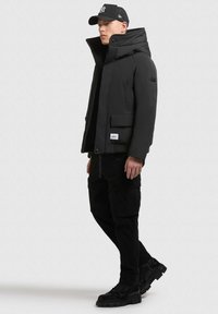 Juodas pūkinis švarkas su aukštu apykakle, priekinėmis kišenėmis ir firminiu ženkleliu, derinamas su juodomis kargo kelnėmis ir batais, užbaigtas juoda kepuraite.