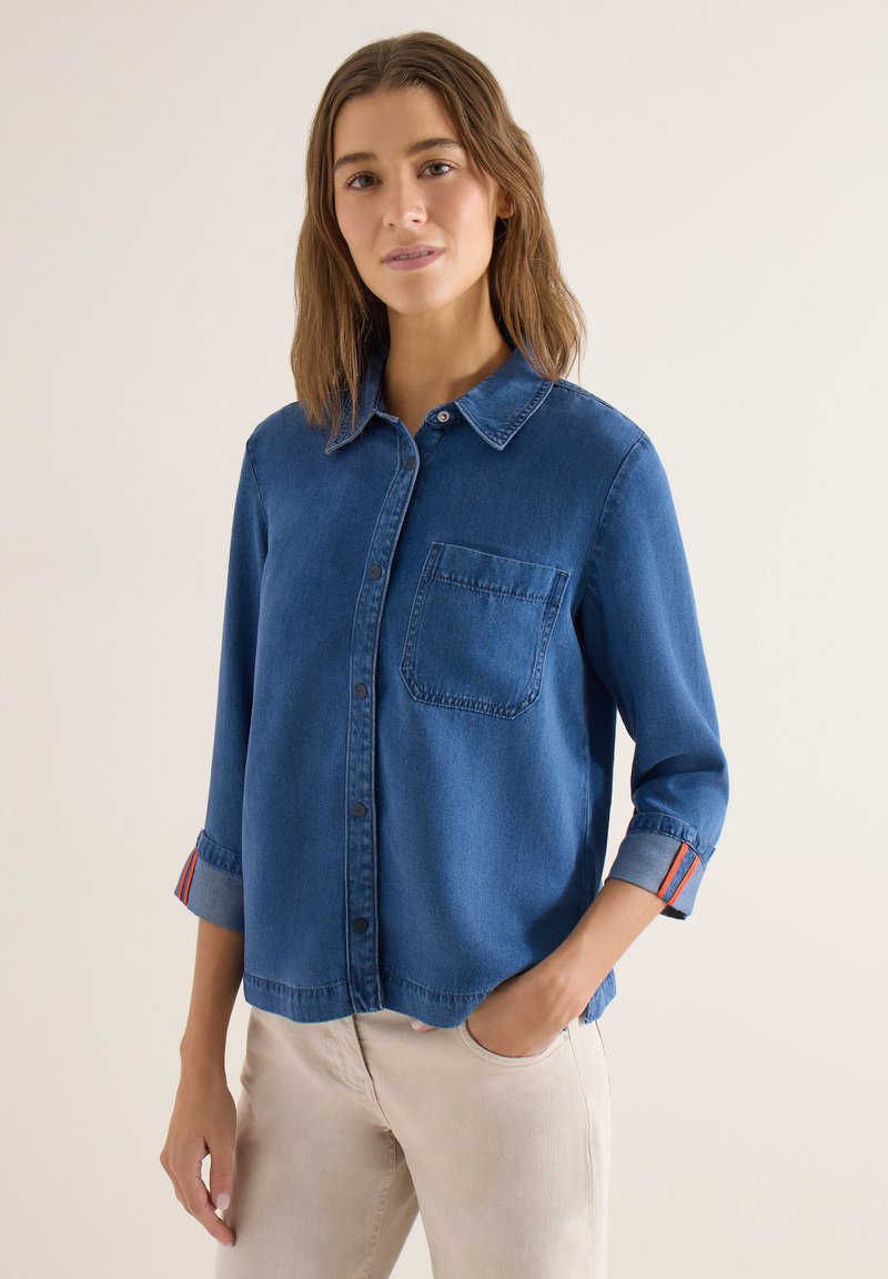 Jeune femme portant une chemise en denim bleue à boutons avec les manches retroussées et un pantalon beige, debout avec une main dans la poche.