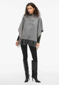 Sieviete ar cirtainiem matiem, tērpusies pelēkā franjā pončo, melnās legingās un melnās augstpapēžu zābaku līdz ceļgaliem, stāv uz balta fona.