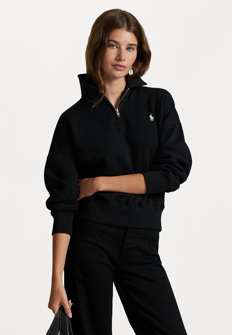Svart quarter-zip sweatshirt med høy krage, ribbede mansjetter og en liten brodert logo på venstre bryst, laget av mykt stoff.